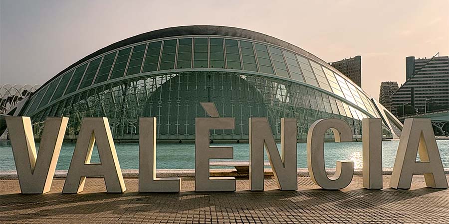 Large “VALENCIA” letters in front of the futuristic glass-covered L’Hemisfèric building, with a reflecting pool and city buildings in the background 
